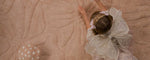 A child wearing a white lace fairy costume lies on a washable wool non-toxic children's rug.