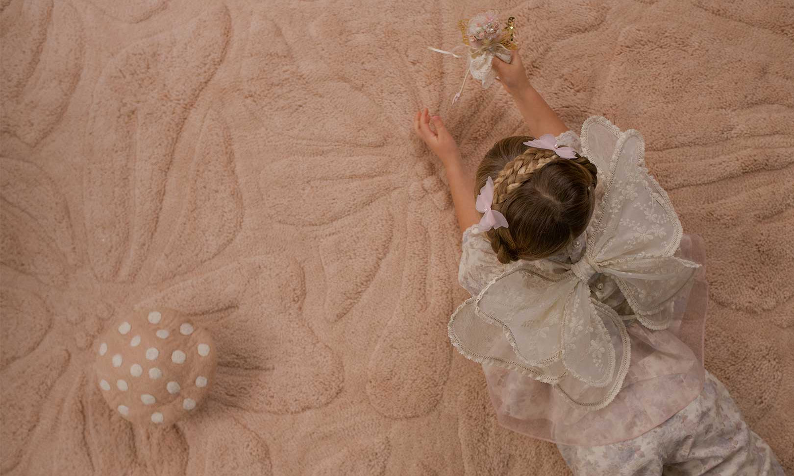 A child wearing a white lace fairy costume lies on a washable wool non-toxic children's rug.