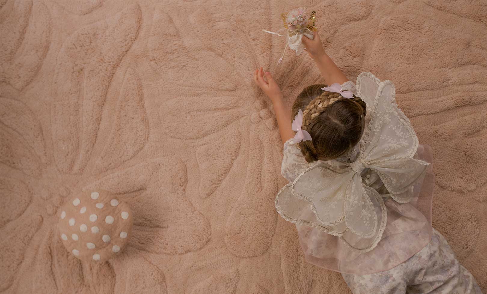 A child wearing a white lace fairy costume lies on a washable wool non-toxic children's rug.